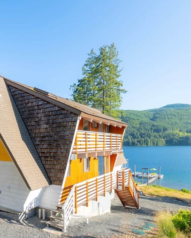 Modern A-Frame cabin with yellow and white exterior on lakeside, surrounded by evergreen trees and mountains