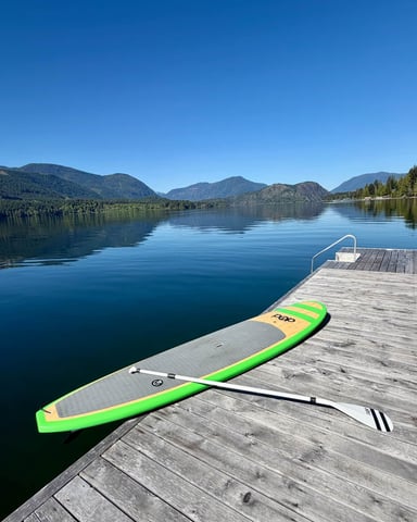 Wooden dock with green paddleboard and paddle on calm alpine lake surrounded by forested mountains