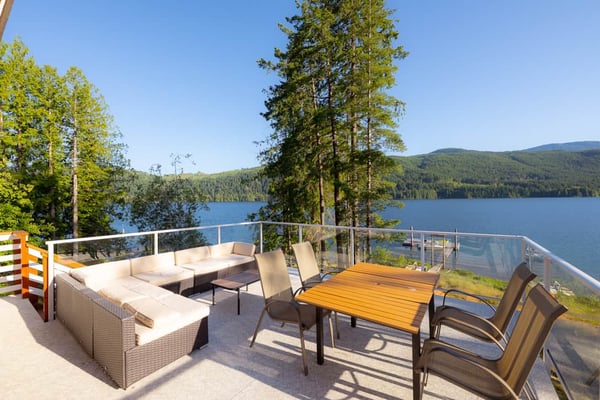 Cabin interior with wooden table, books, glasses, and mug overlooking forested lake with dock