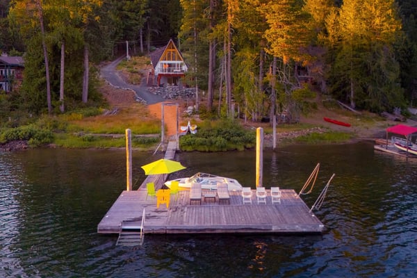 Modern dark cabin with wooden deck and outdoor seating overlooking forested lake at sunset