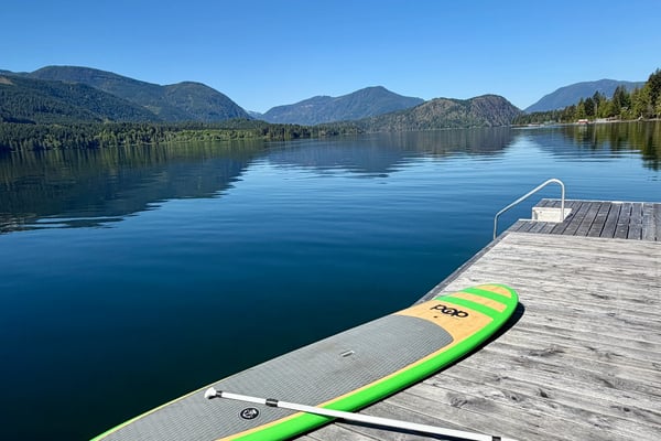 Paddleboard and paddle on wooden dock overlooking calm alpine lake with forested mountains