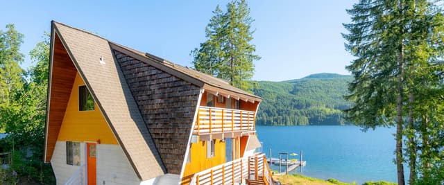 Lakefront dock with yellow umbrella and chairs, A-Frame cabin in forest with autumn foliage, boats moored nearby