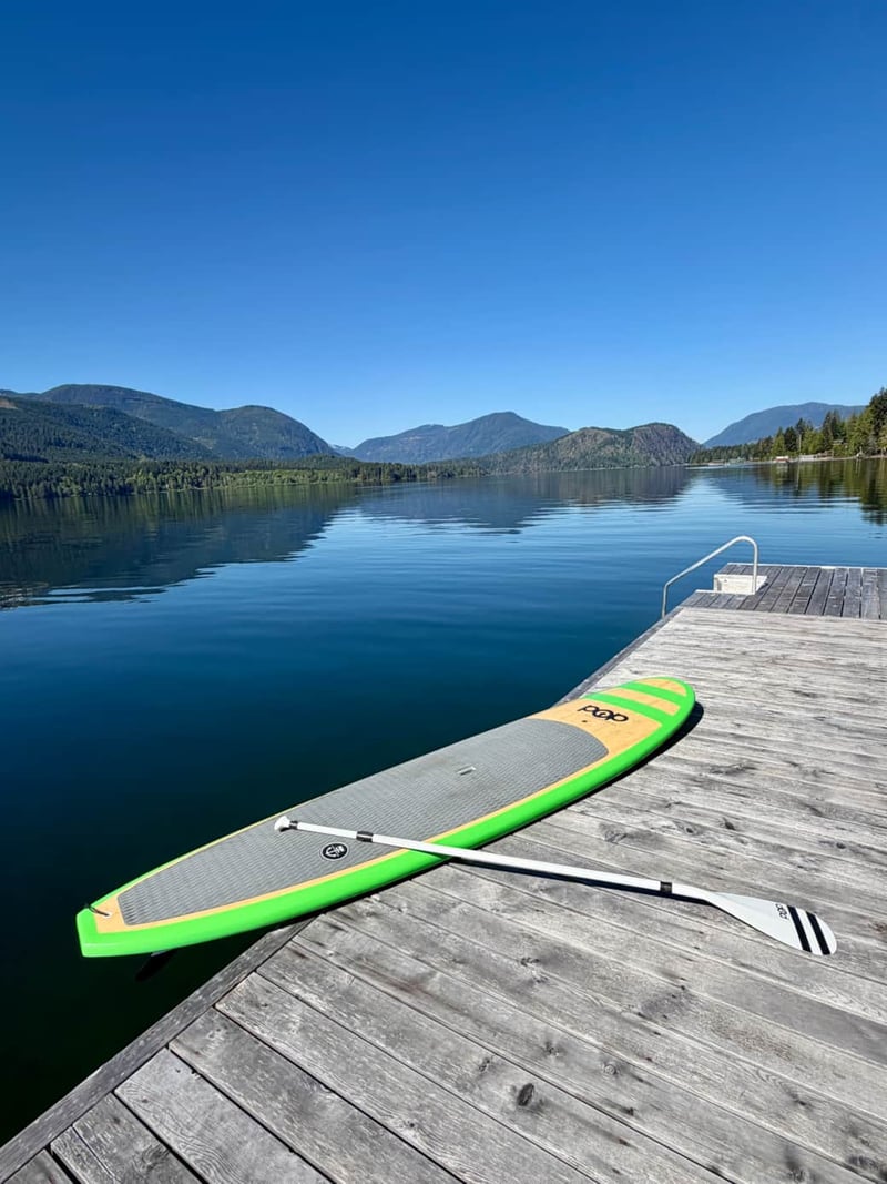 Paddleboard and paddle on wooden dock overlooking calm alpine lake surrounded by forested mountains