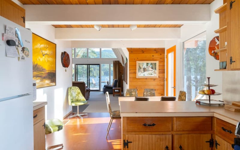 Open-plan kitchen and living area with wood beams, large windows overlooking forest, and lake view through glass doors
