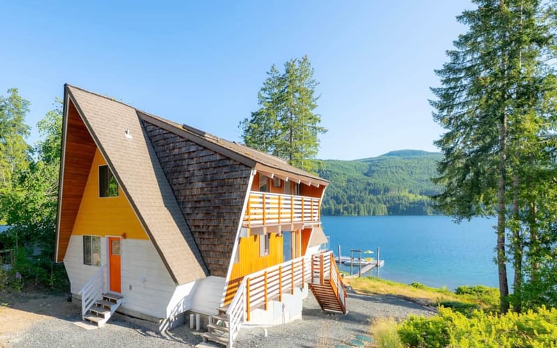 Modern A-Frame cabin with yellow and white exterior, dark roof, and wooden decks overlooking a blue lake surrounded by evergreen forest.