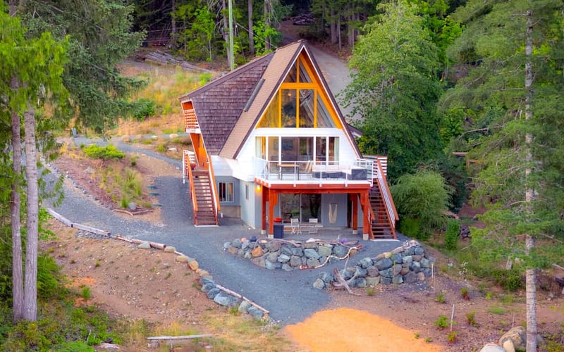 Modern A-Frame cabin with red and white exterior surrounded by dense forest and gravel driveway