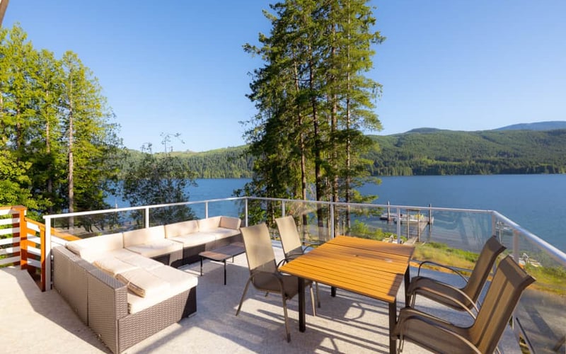 Waterfront deck with lounge seating, dining table, and glass railing overlooking forested lake and mountains