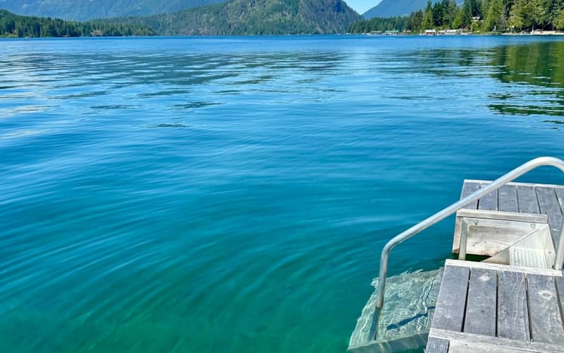 Wooden dock with metal railing extending into clear blue alpine lake, forested mountains in background